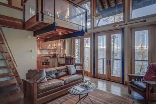 Cozy living room with wooden furniture, large windows overlooking a lake, and a lofted area above.